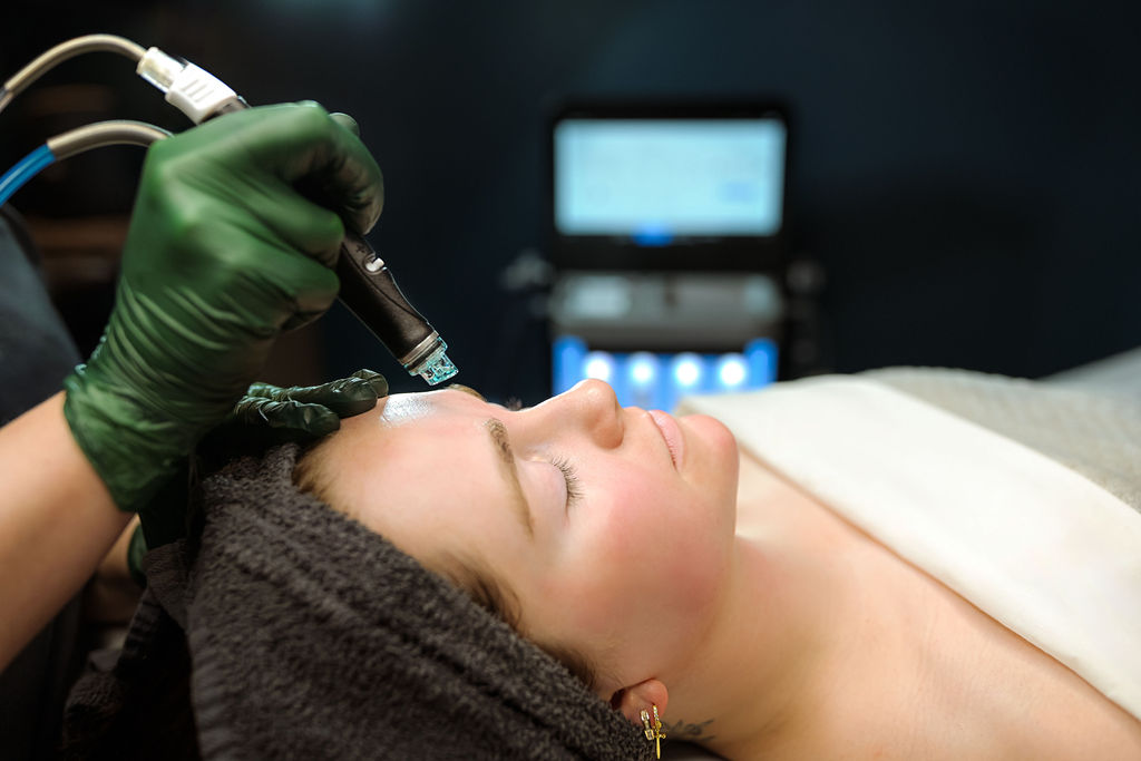 Person receiving a cosmetic treatment on forehead in a clinical setting, with medical equipment in background.