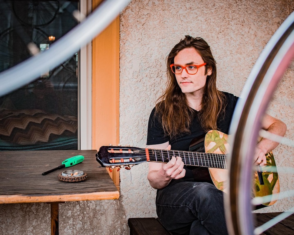 Woman with long hair and red glasses playing guitar outdoors near a wall and window.