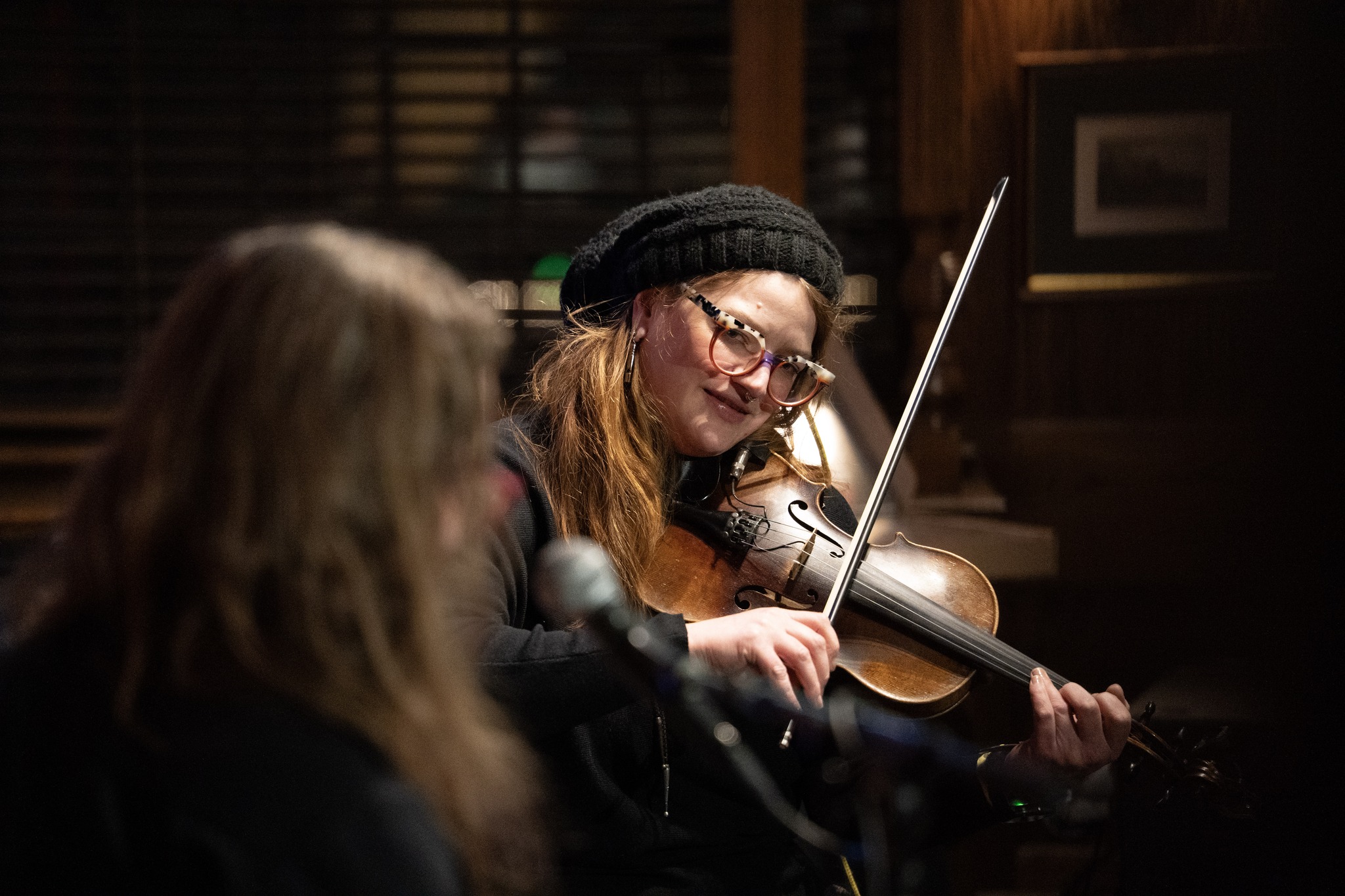 Woman playing violin, smiling, wearing glasses and a black beanie, in a dimly lit room with wooden walls.