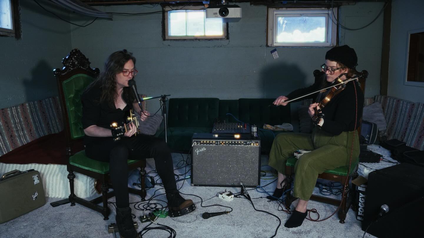 Two women playing guitars in a room with a small amplifier between them, sitting on chairs, with a sofa and windows in background.