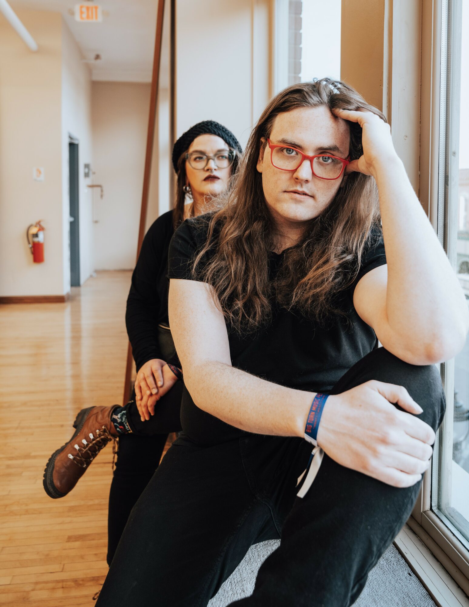 Two women sitting indoors near a window, one in the foreground with long hair and glasses, the other in the background with glasses.