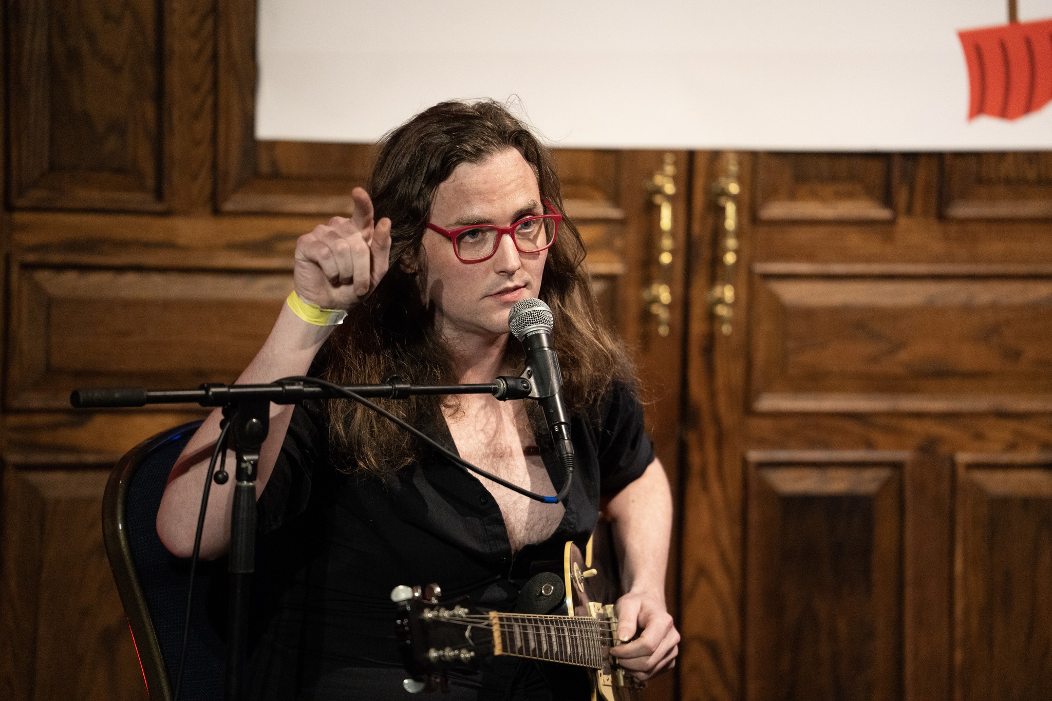 Woman with glasses and dark hair speaking into a microphone, gesturing with her right hand, in a wood-paneled room.