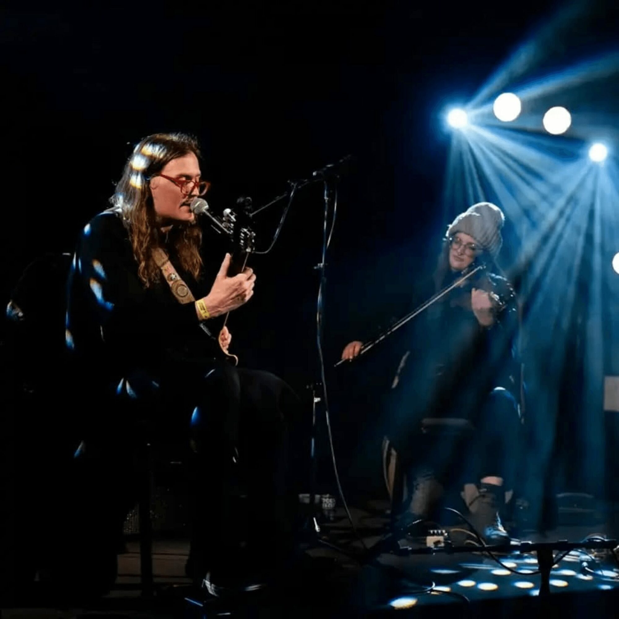 Two women perform on stage with microphones and musical instruments, illuminated by stage lights, dark background.