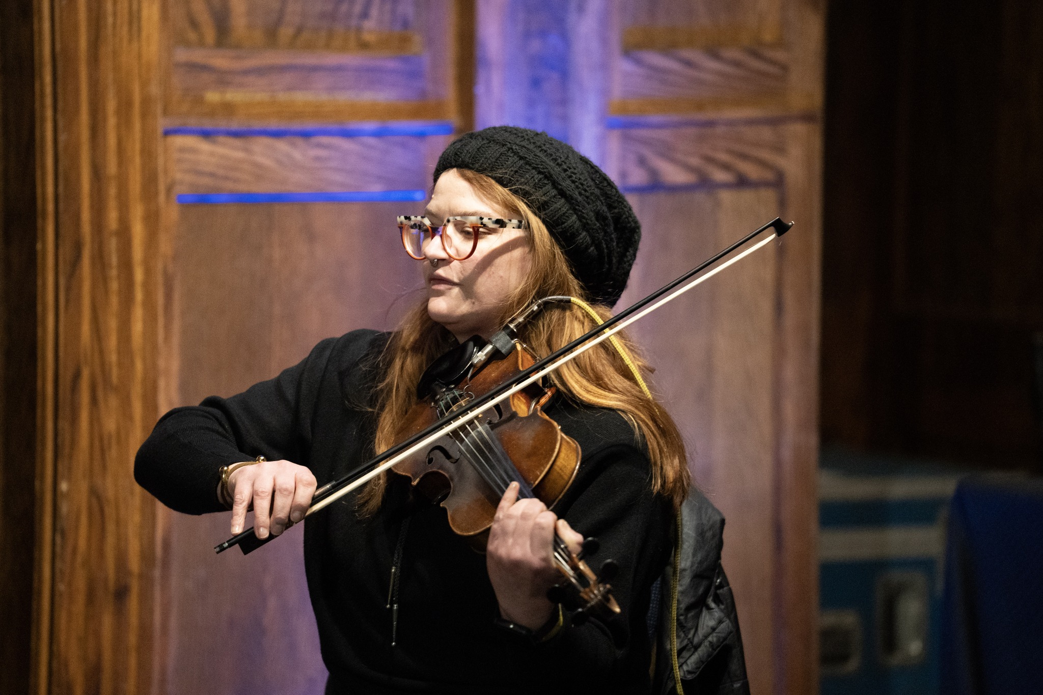 Woman playing violin, wearing glasses, black beanie, and black jacket, in a wooden-paneled room.