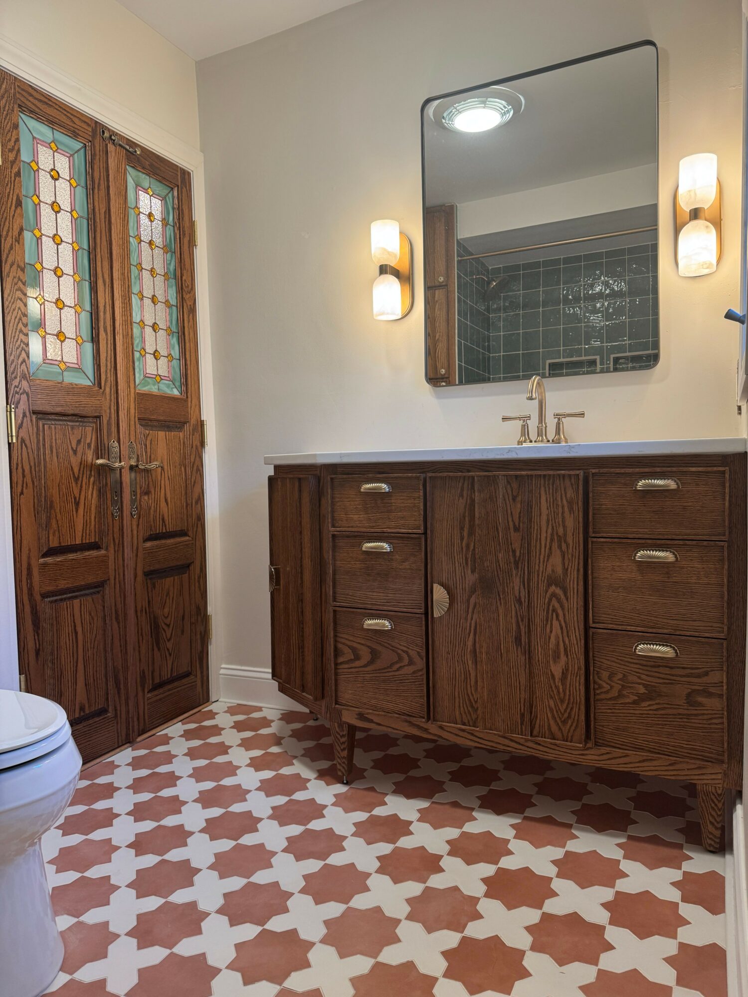 Bathroom vanity with mirror, two wall sconces, wooden cabinet, and patterned tile floor.