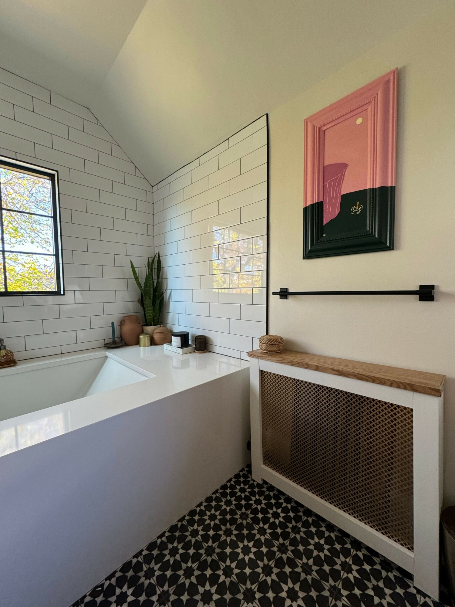 Bathroom corner with white bathtub, window, potted plant, framed pink artwork, and patterned black and white floor.