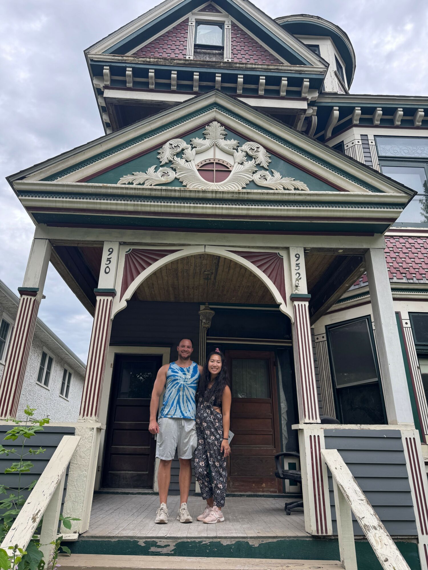 Two people standing on porch in front of large Victorian-style house with decorative trim and gabled roof.