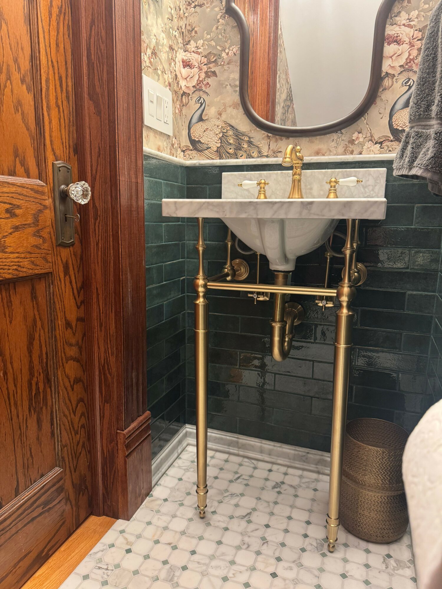 Bathroom sink with gold fixtures, supported by gold legs, in front of green tiled wall and mirror.
