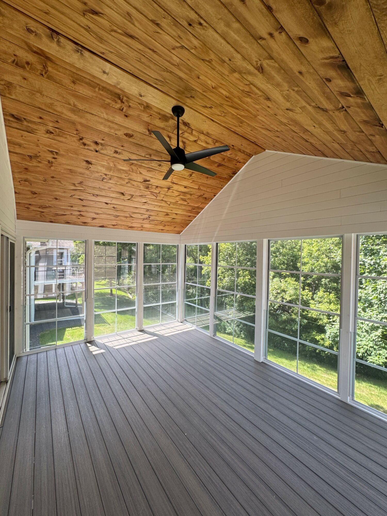 Sunroom with wooden ceiling, ceiling fan, large windows, and gray flooring, overlooking green outdoor scenery.