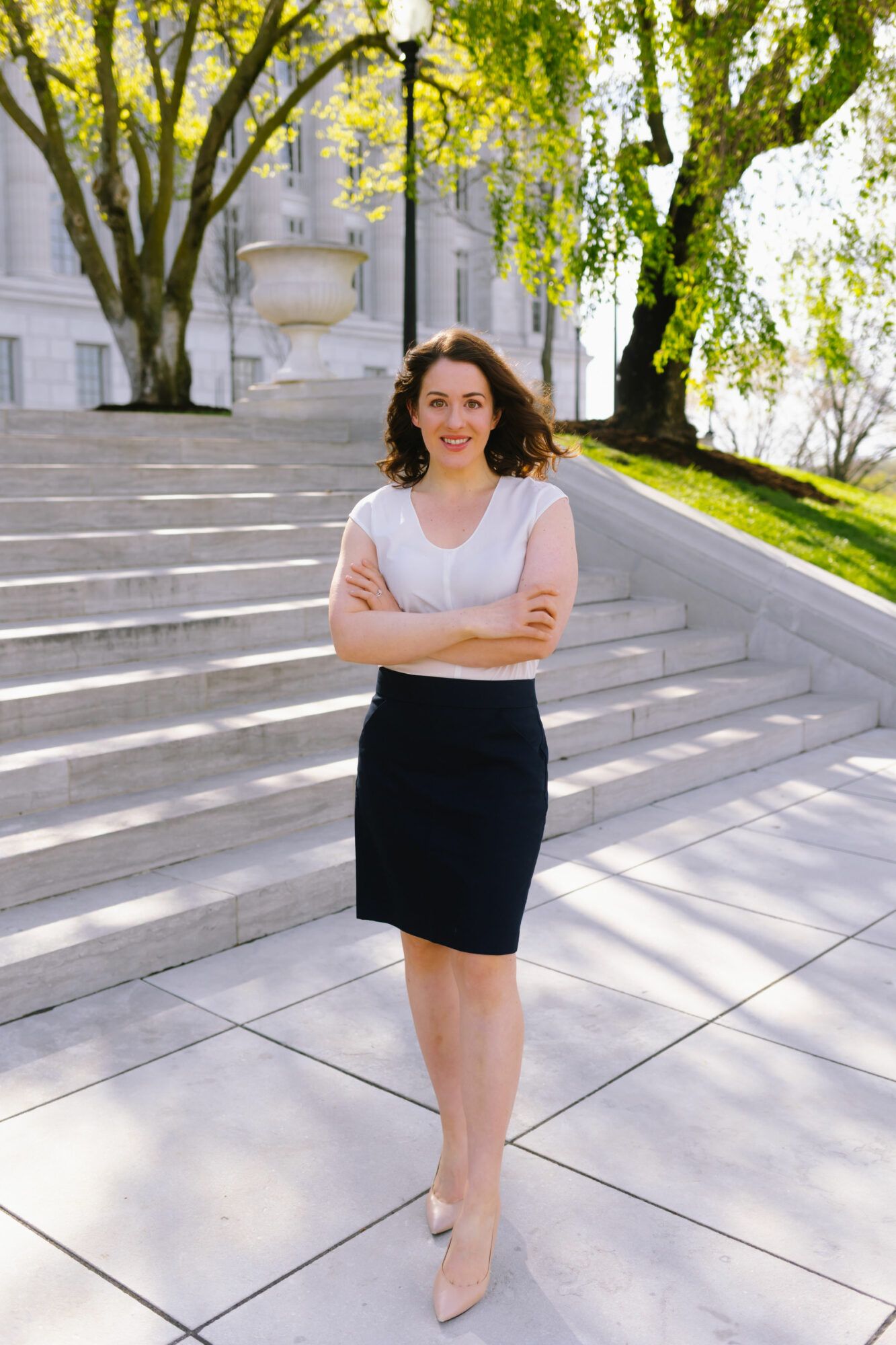 Woman standing outdoors on stairs with trees and building in background, smiling, arms crossed, wearing white top and black skirt.