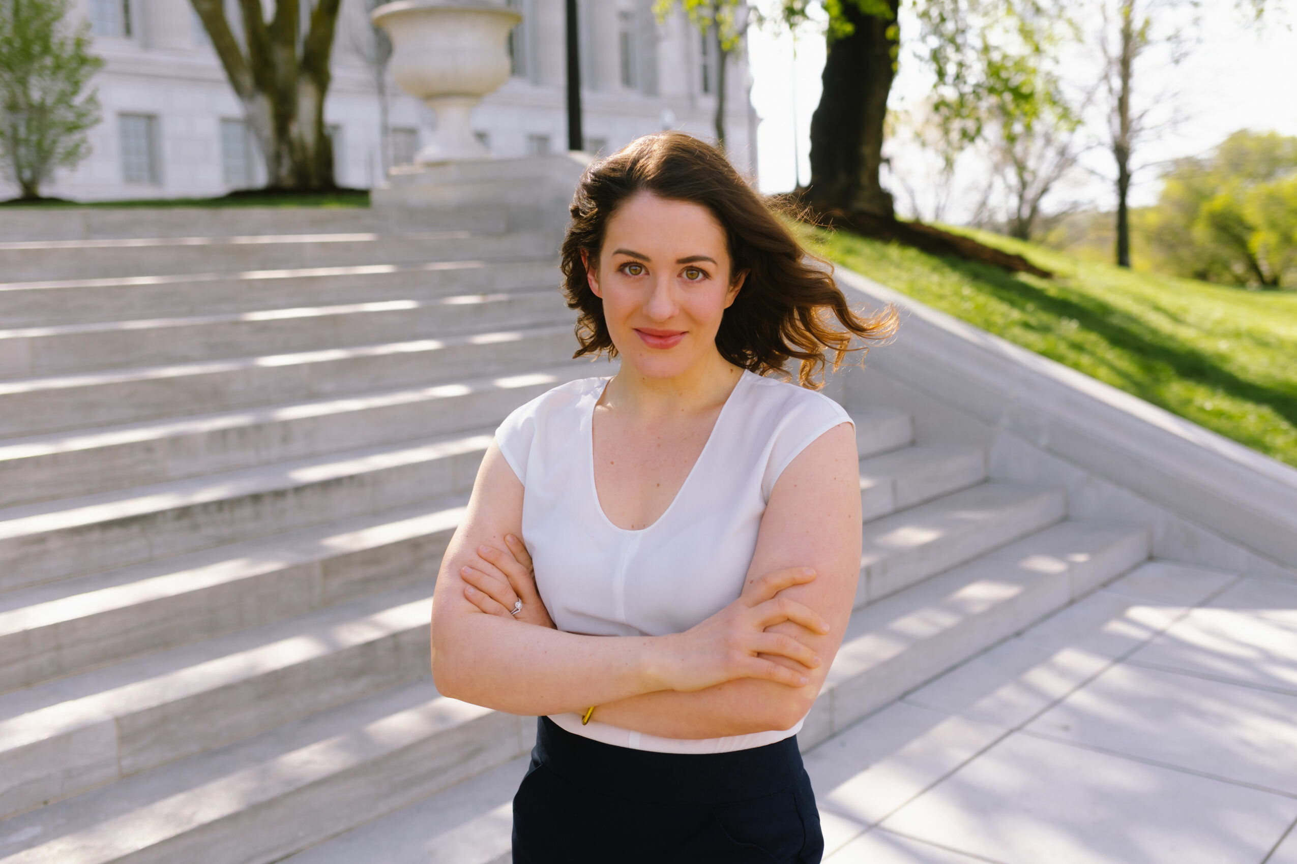 Woman with shoulder-length curly hair standing outdoors on steps, smiling, arms crossed, in sunlight.
