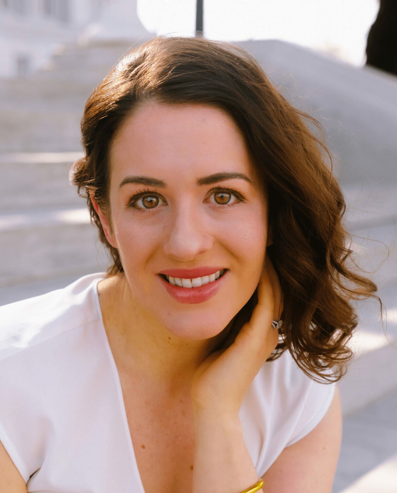 Woman with shoulder-length wavy brown hair, smiling, wearing a white top, outdoors with stairs in background.