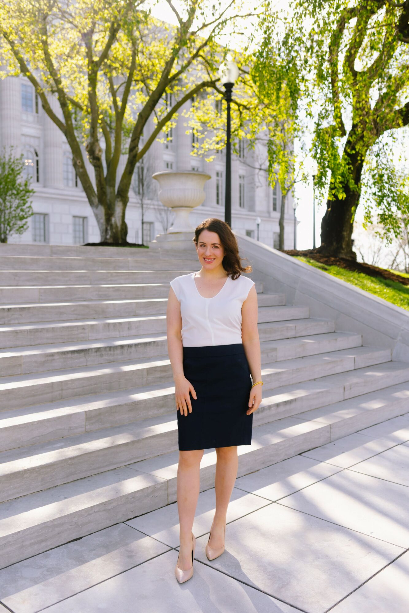 Woman in white top and black skirt standing on steps outdoors with trees and building in background.