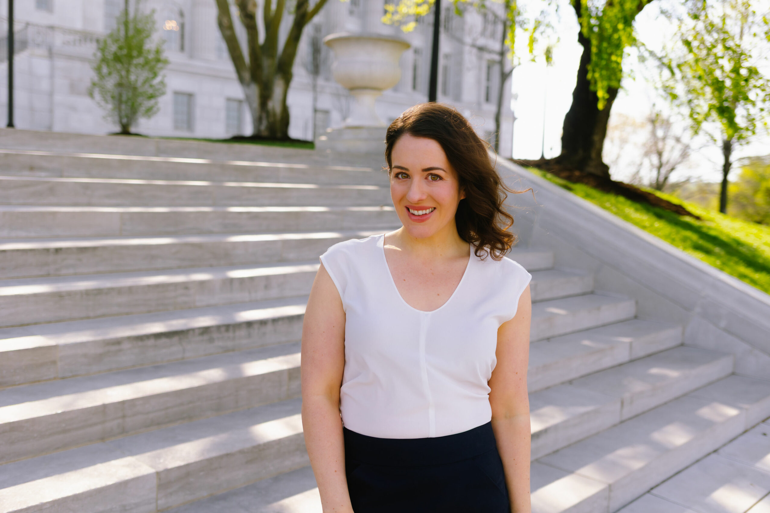 Woman with dark hair in a white top and dark skirt standing on outdoor stairs, smiling.