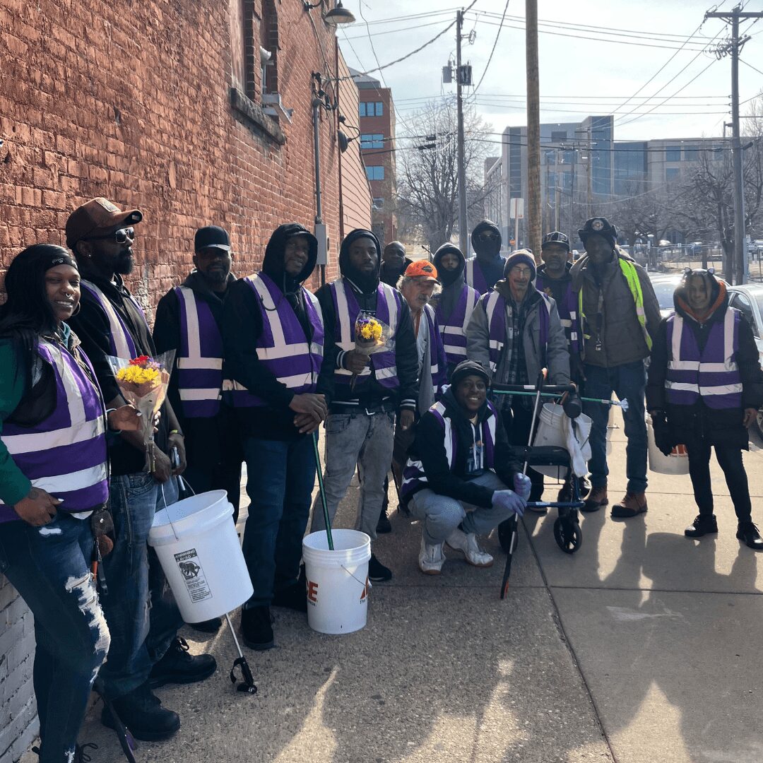 Group of people wearing purple reflective vests standing on sidewalk near brick building, some holding flowers and buckets.