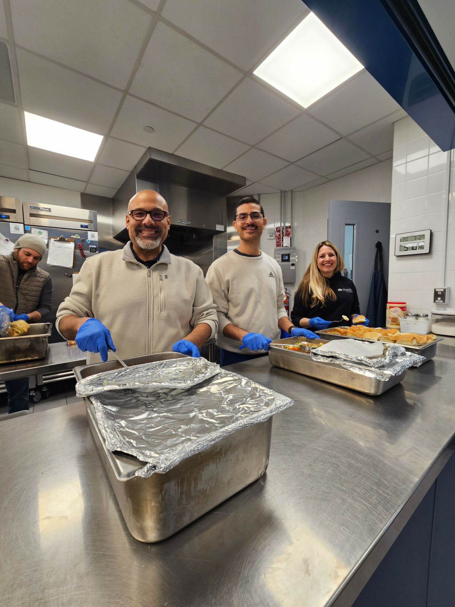Three people in a kitchen, smiling, wearing gloves, with trays of food covered in foil on a stainless steel counter.