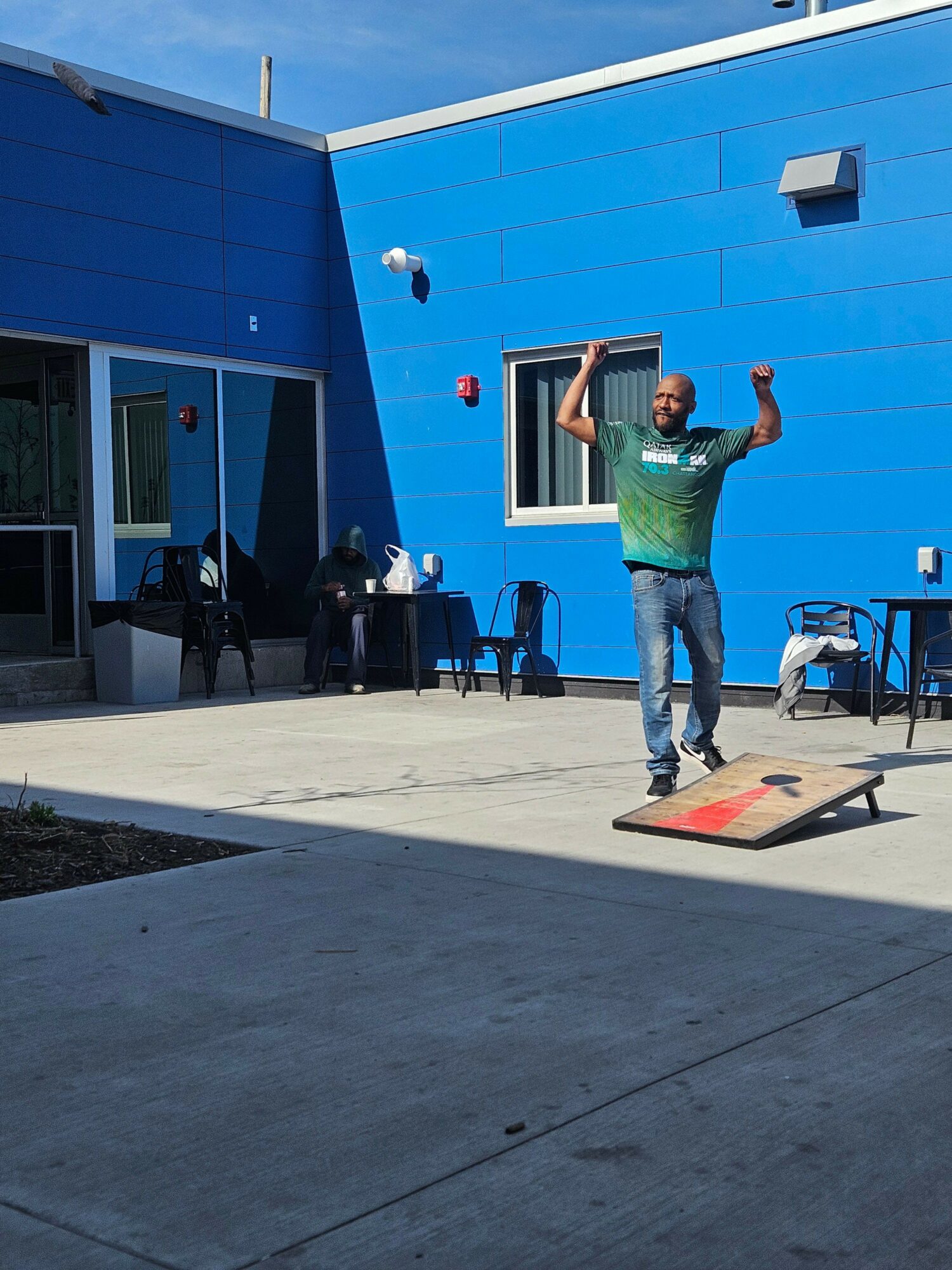 Person playing cornhole outdoors near a blue building, throwing a bean bag with a blue sky background.