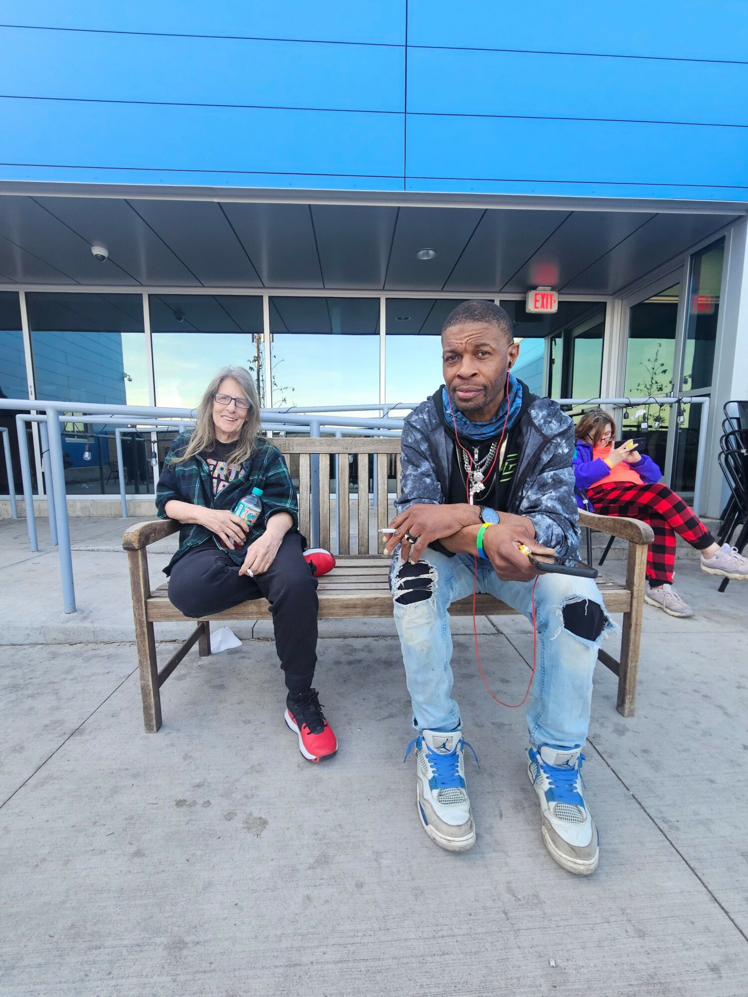 Two people sitting on a bench outside a building with large glass windows, one wearing glasses and the other in a jacket.
