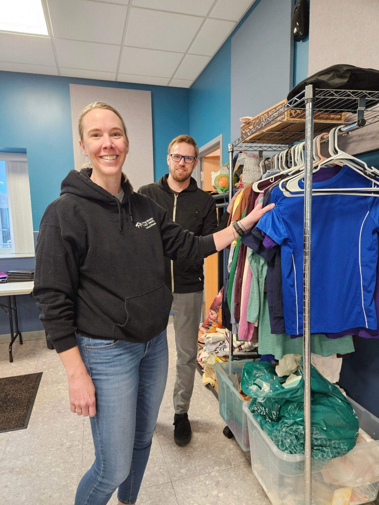 Two people in a room with blue walls, one woman smiling and touching clothing on a rack, a man in the background.