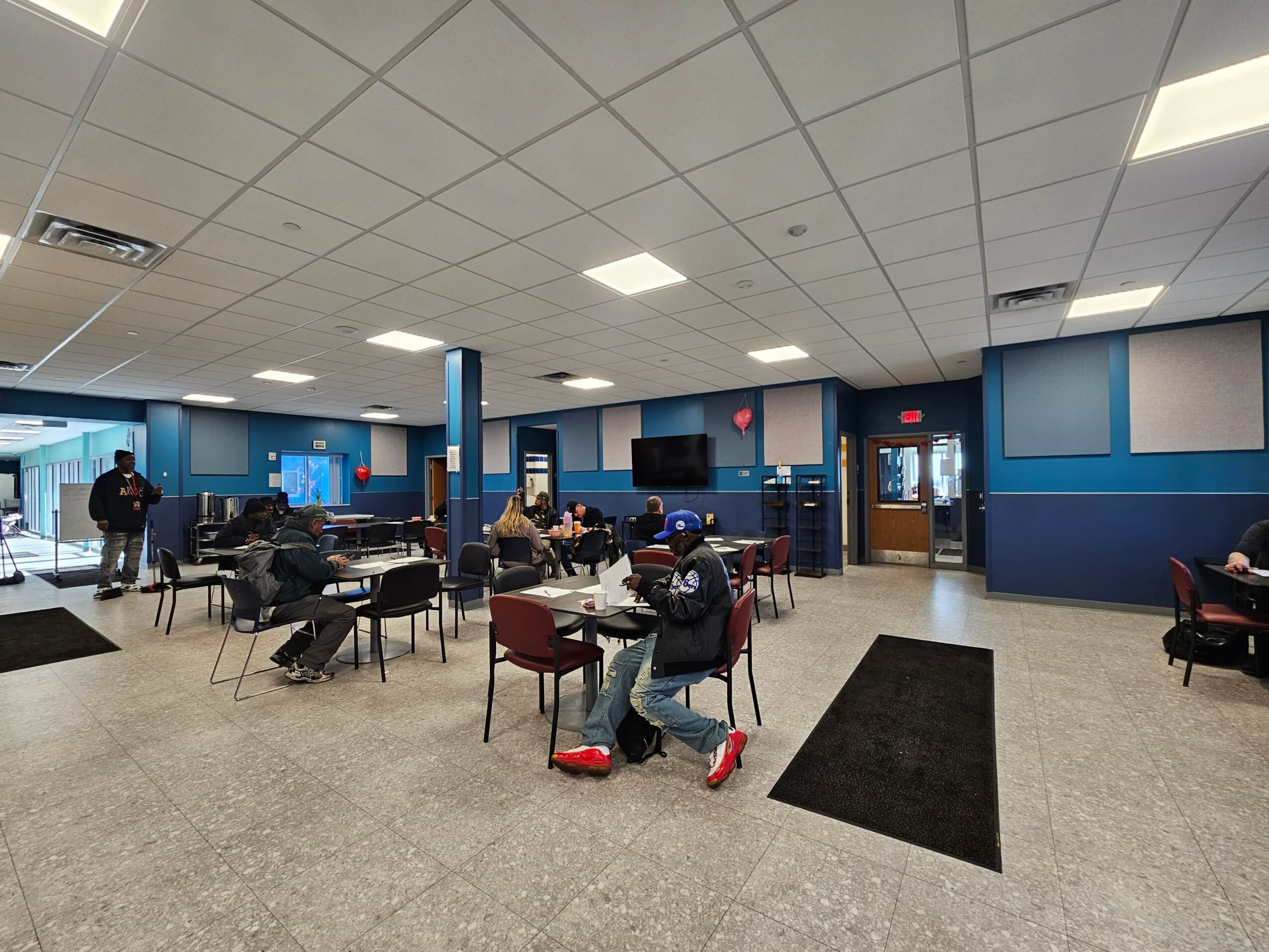 People sitting and standing in a spacious room with blue walls, tables, chairs, and a large screen on the wall.