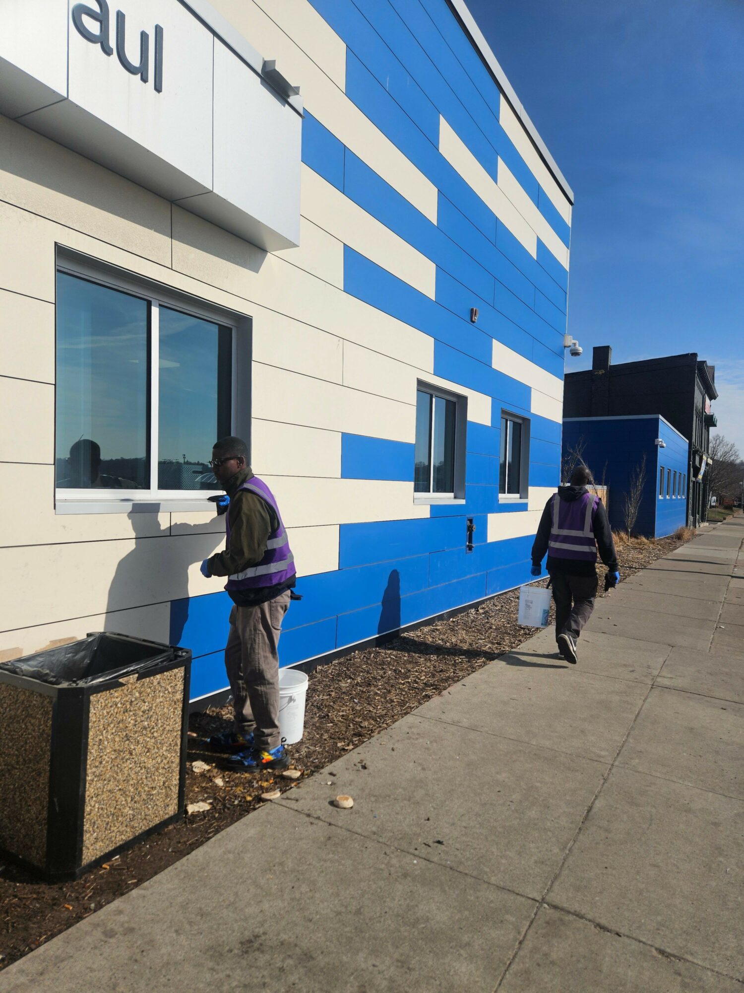 Two people walking past a building with a blue and white geometric pattern exterior, windows, and a sidewalk.