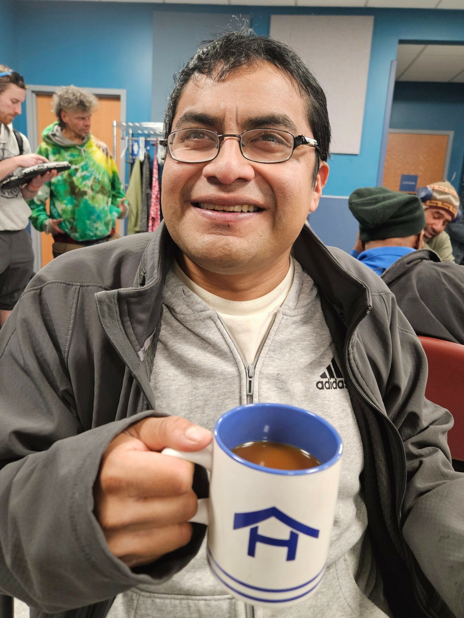 Smiling man holding a mug with a blue logo, in a busy indoor setting with people in the background.