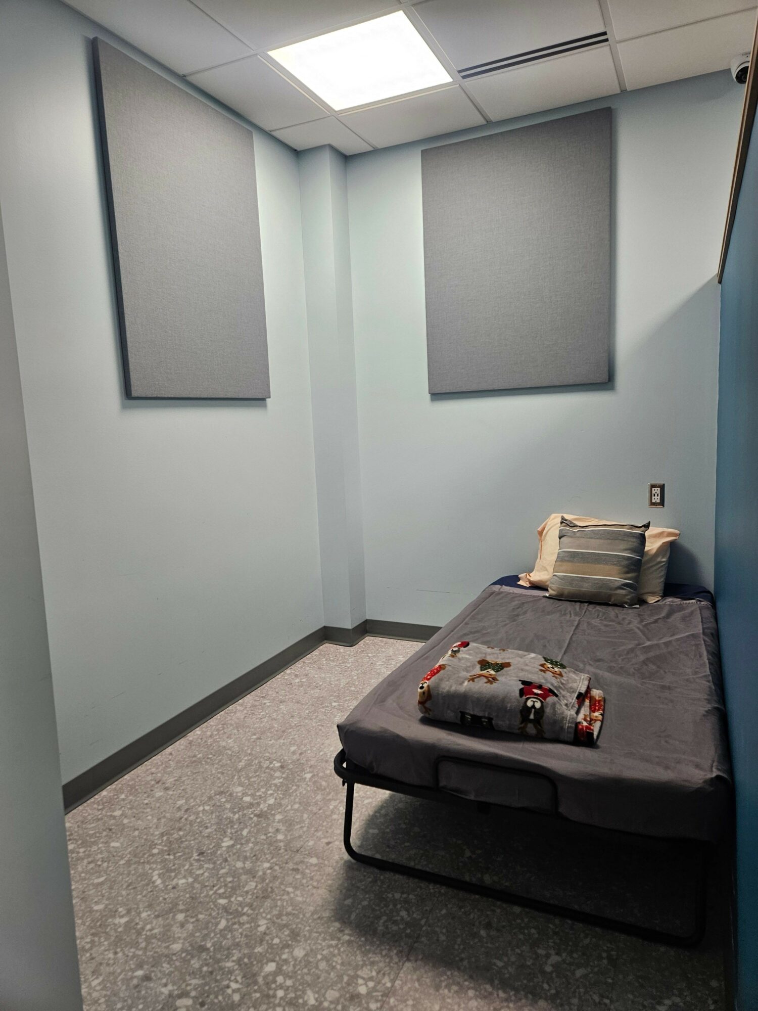Small room with gray acoustic panels on walls, single bed with pillow and blanket, plain walls and ceiling, tiled floor.