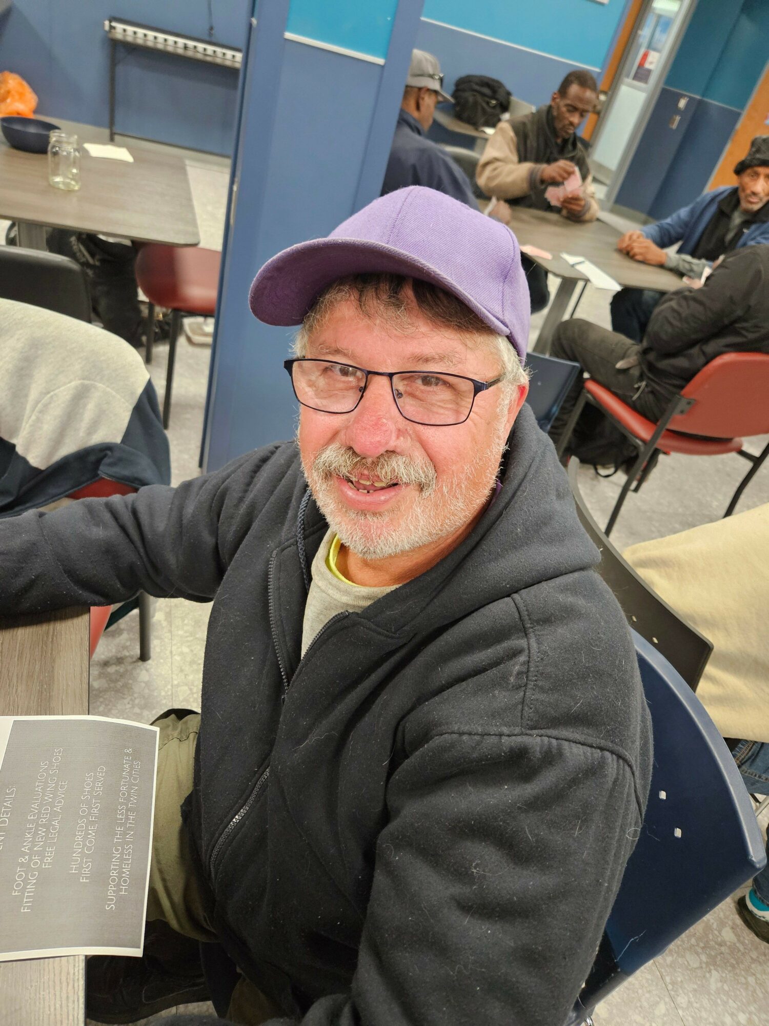 Man with glasses, purple cap, and gray hoodie smiling at the camera in a room with tables and people.