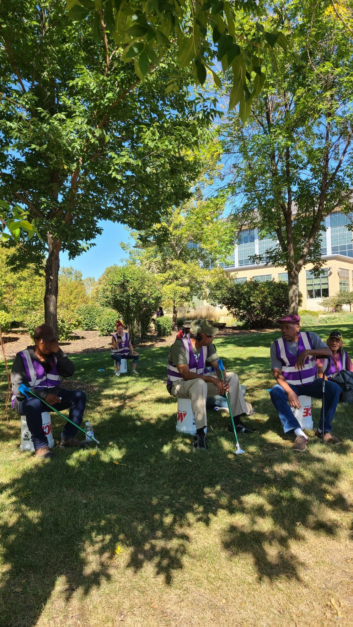 Group of people sitting on chairs outdoors under trees in a park, with buildings in the background.