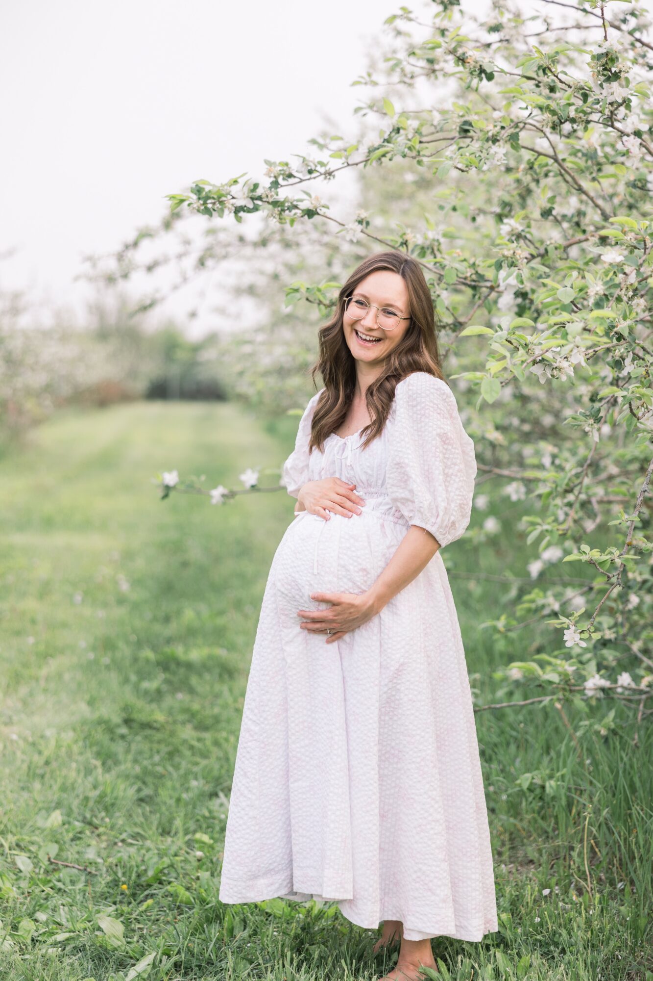 Pregnant woman in a white dress standing outdoors near blooming trees, smiling, holding her belly.