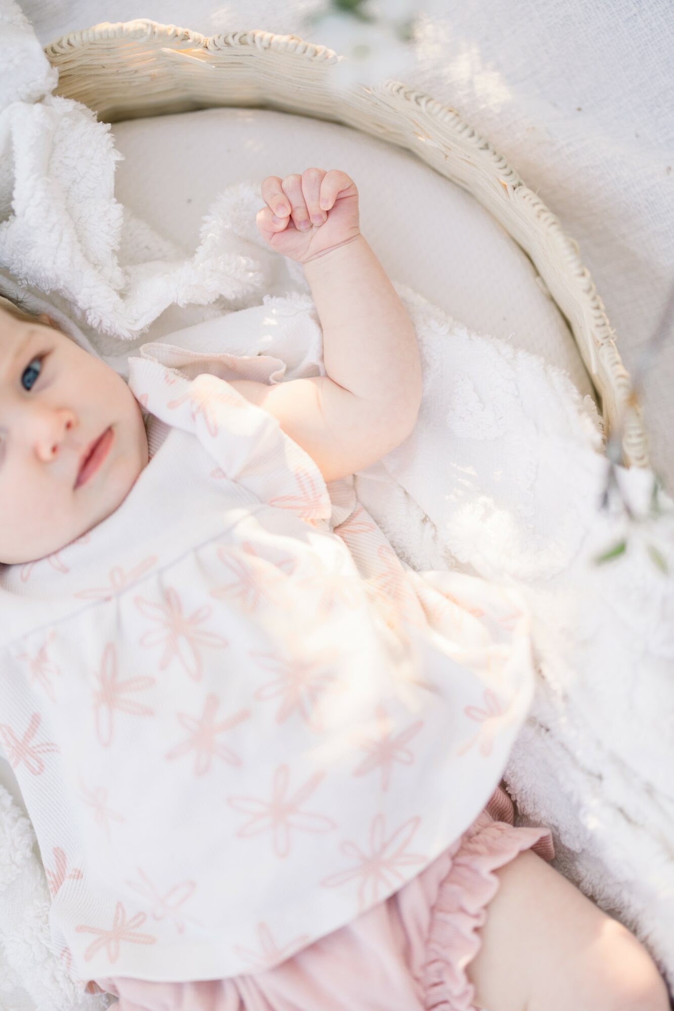 Baby lying in a crib, wearing a white dress with pink patterns, surrounded by soft blankets.