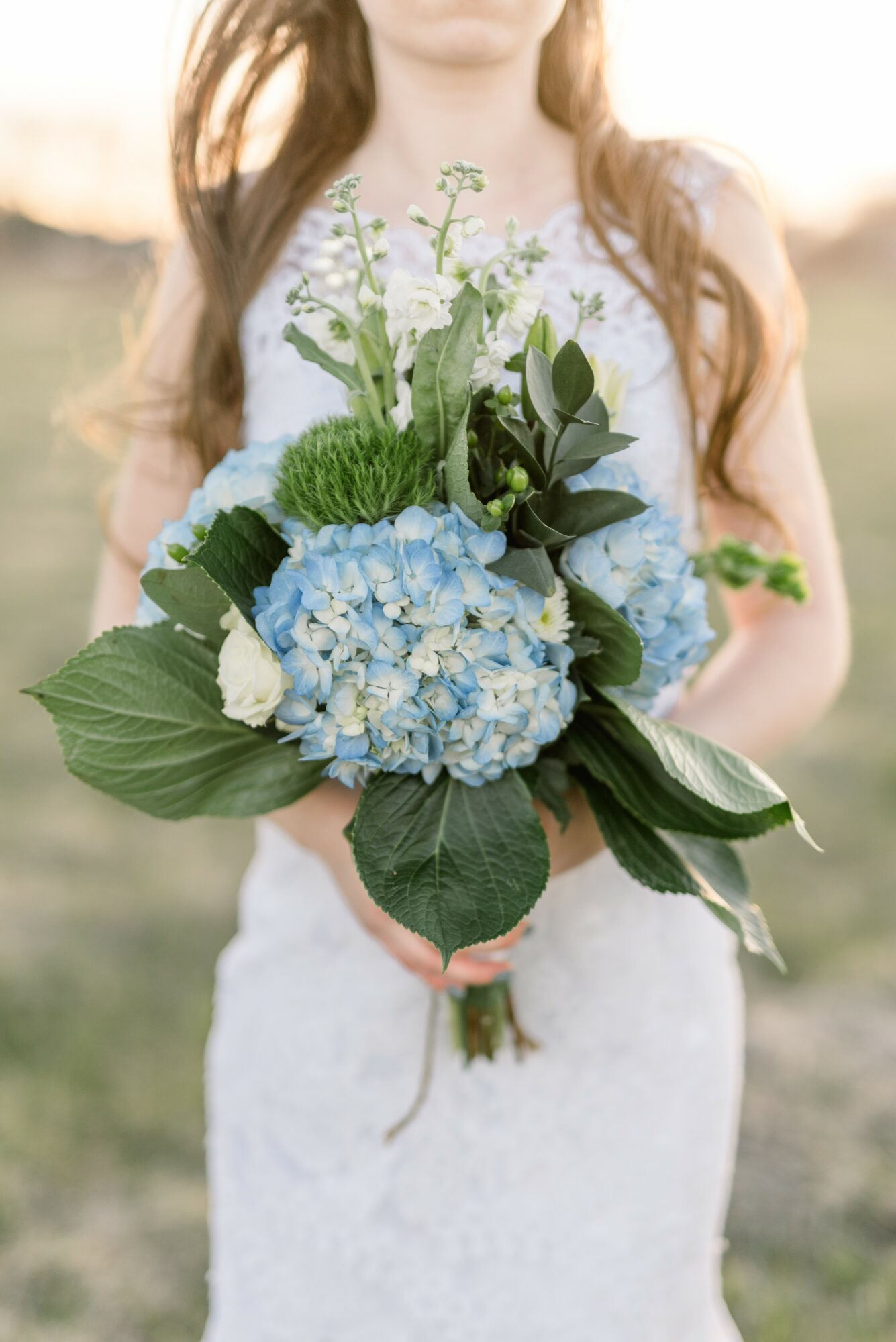 Person holding a bouquet of blue hydrangeas, white roses, and green foliage outdoors.