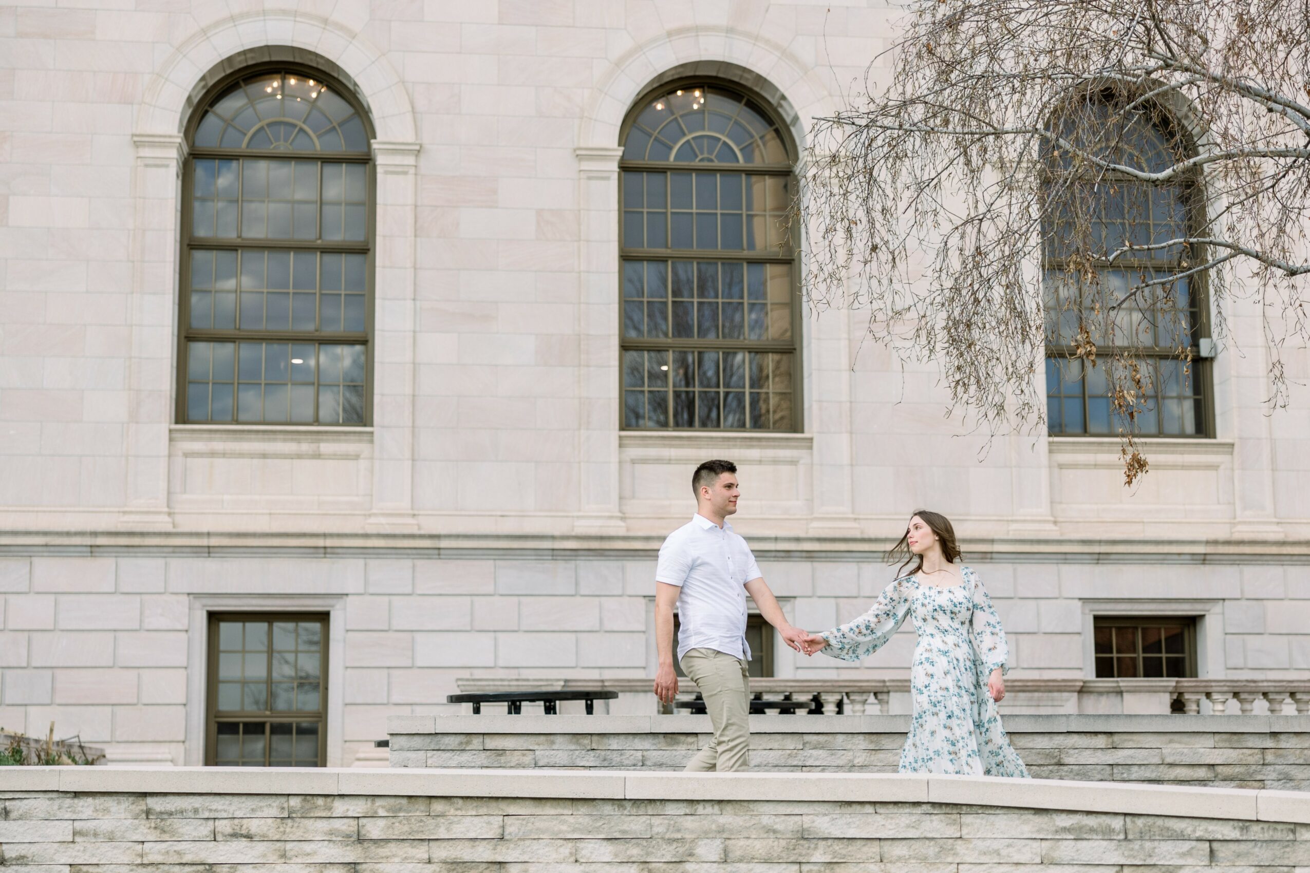 A man and woman holding hands in front of a stone building with arched windows, outdoors during daytime.