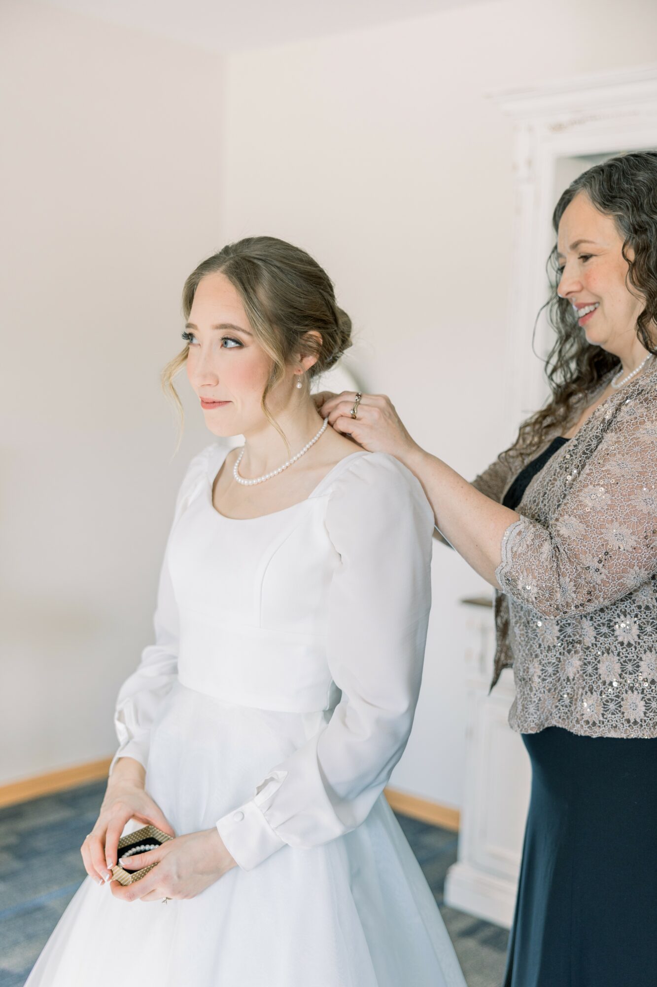 Two women, one in a white dress, the other adjusting her hair, in a bright room with white walls.
