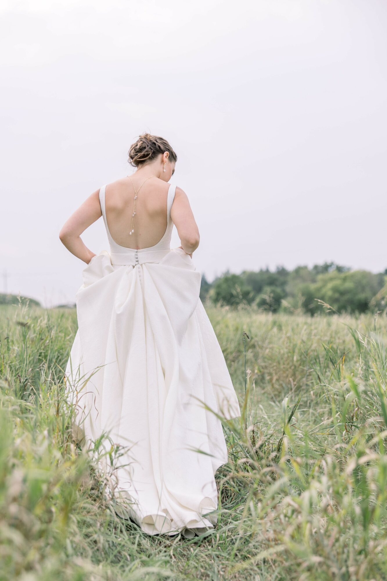 Woman in a white dress walking away in a grassy field, back view, overcast sky, natural landscape.
