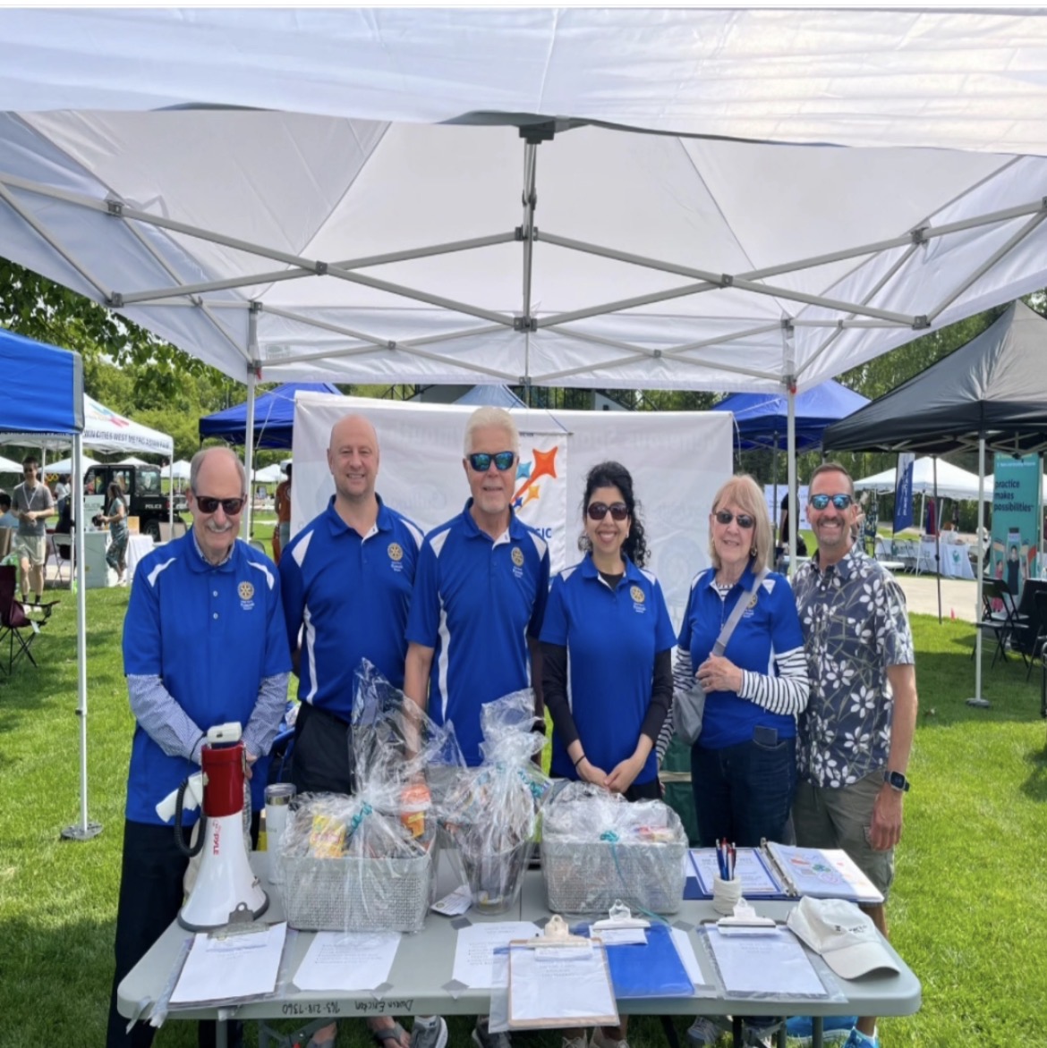Six people standing behind a table with baskets and documents under a white canopy at an outdoor event.
