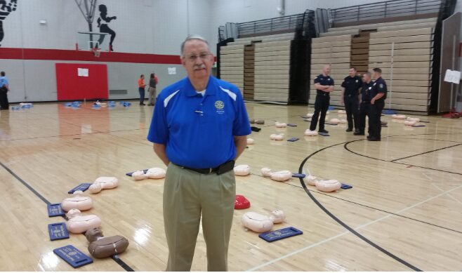 Man in blue shirt and beige pants standing in gymnasium with bowling pins and balls arranged on the floor.