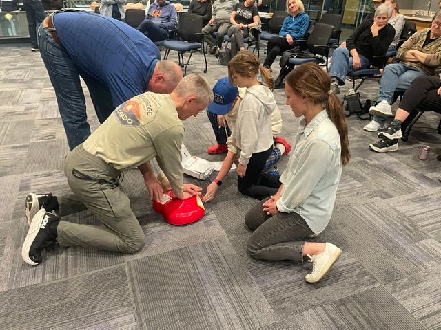 Two adults and two children practicing CPR on a mannequin in a room with seated people.