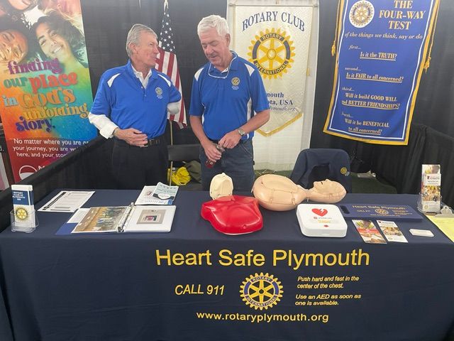 Two men in blue shirts stand behind a table with heart-shaped models, banners, and informational materials at a Heart Safe Plymouth event.