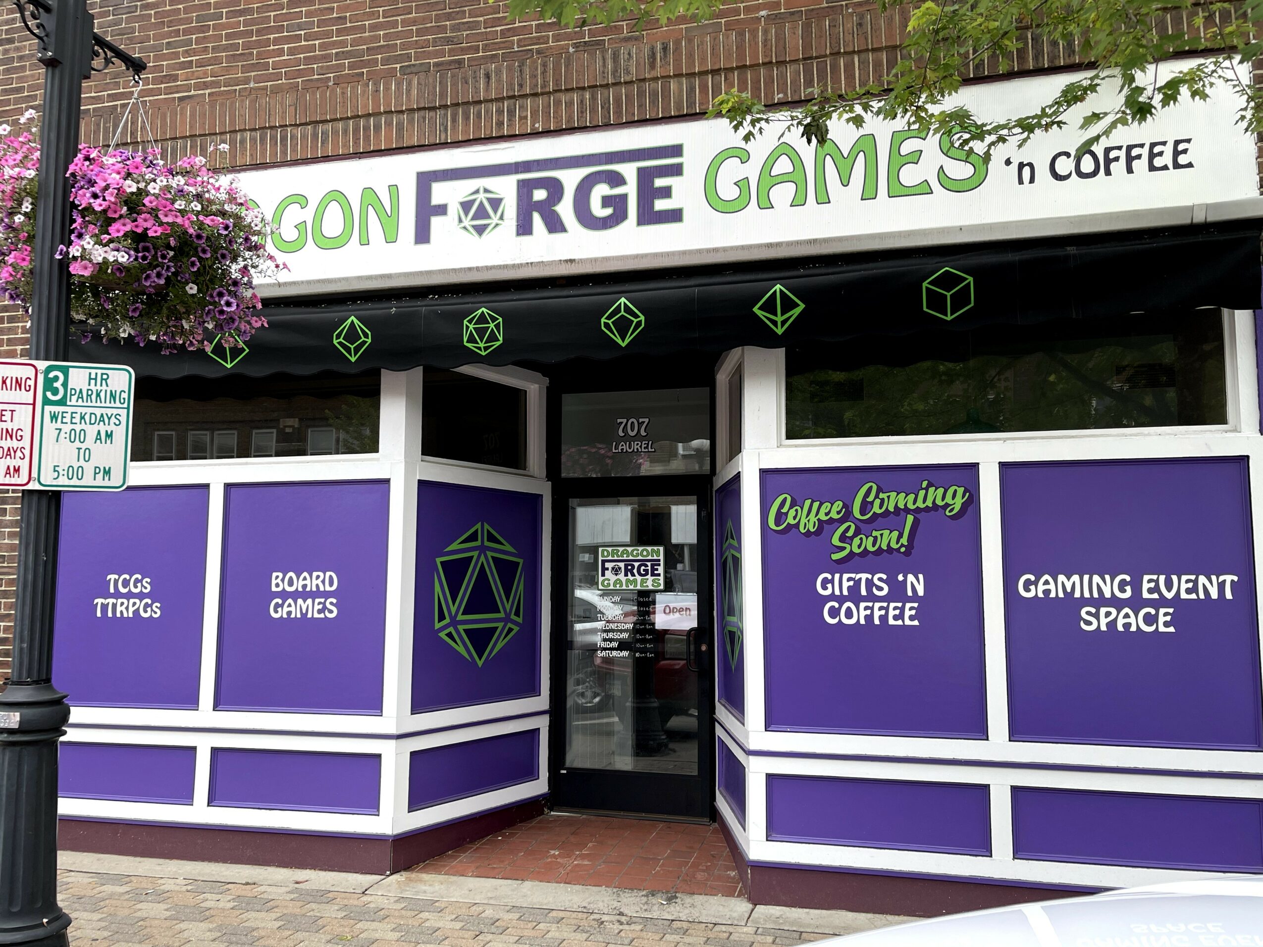Storefront with purple and white signage for board games and coffee, entrance door in center, hanging flower basket on left.