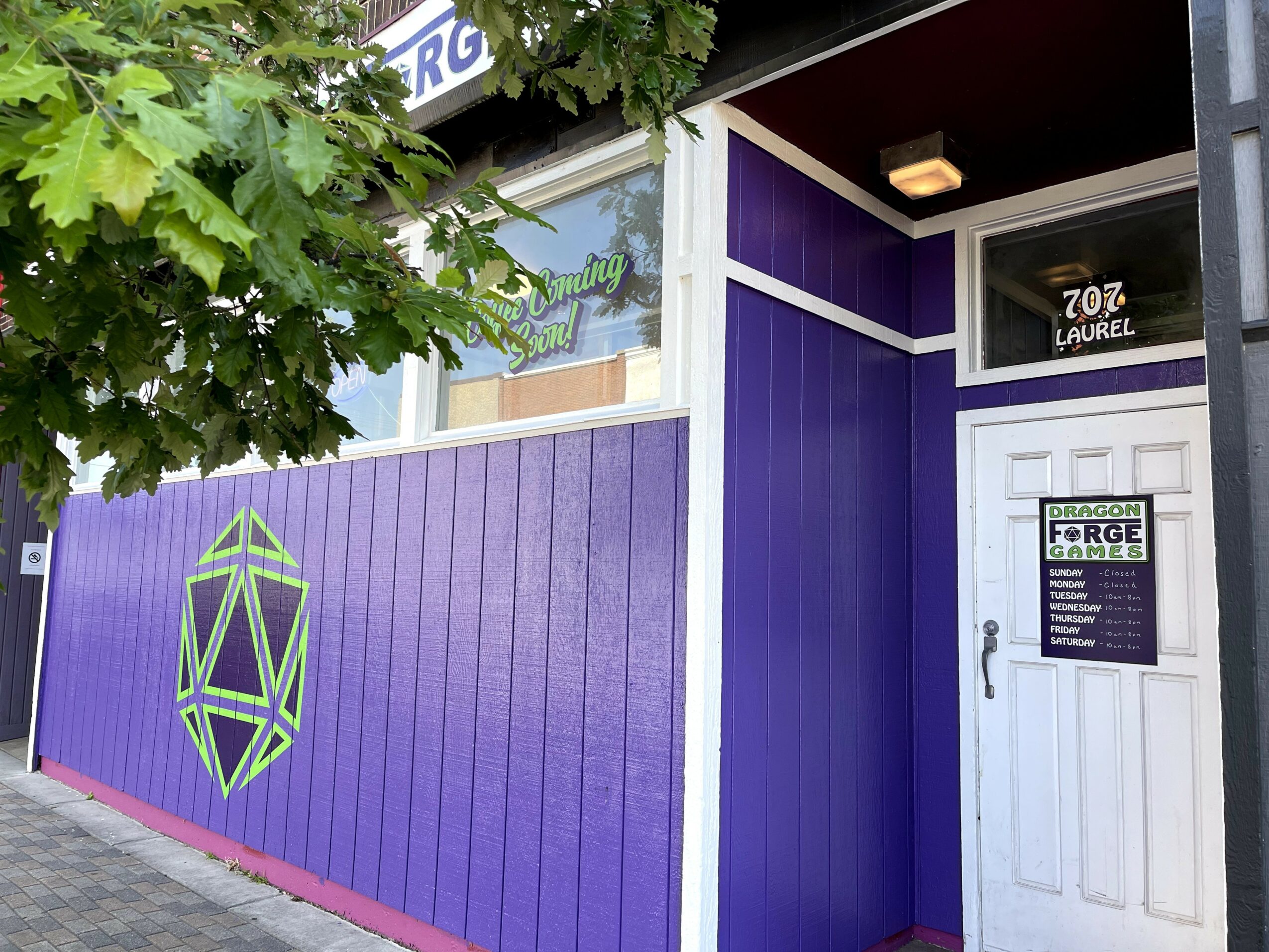 Purple building exterior with geometric logo, white door, and window, partially obscured by green tree leaves.