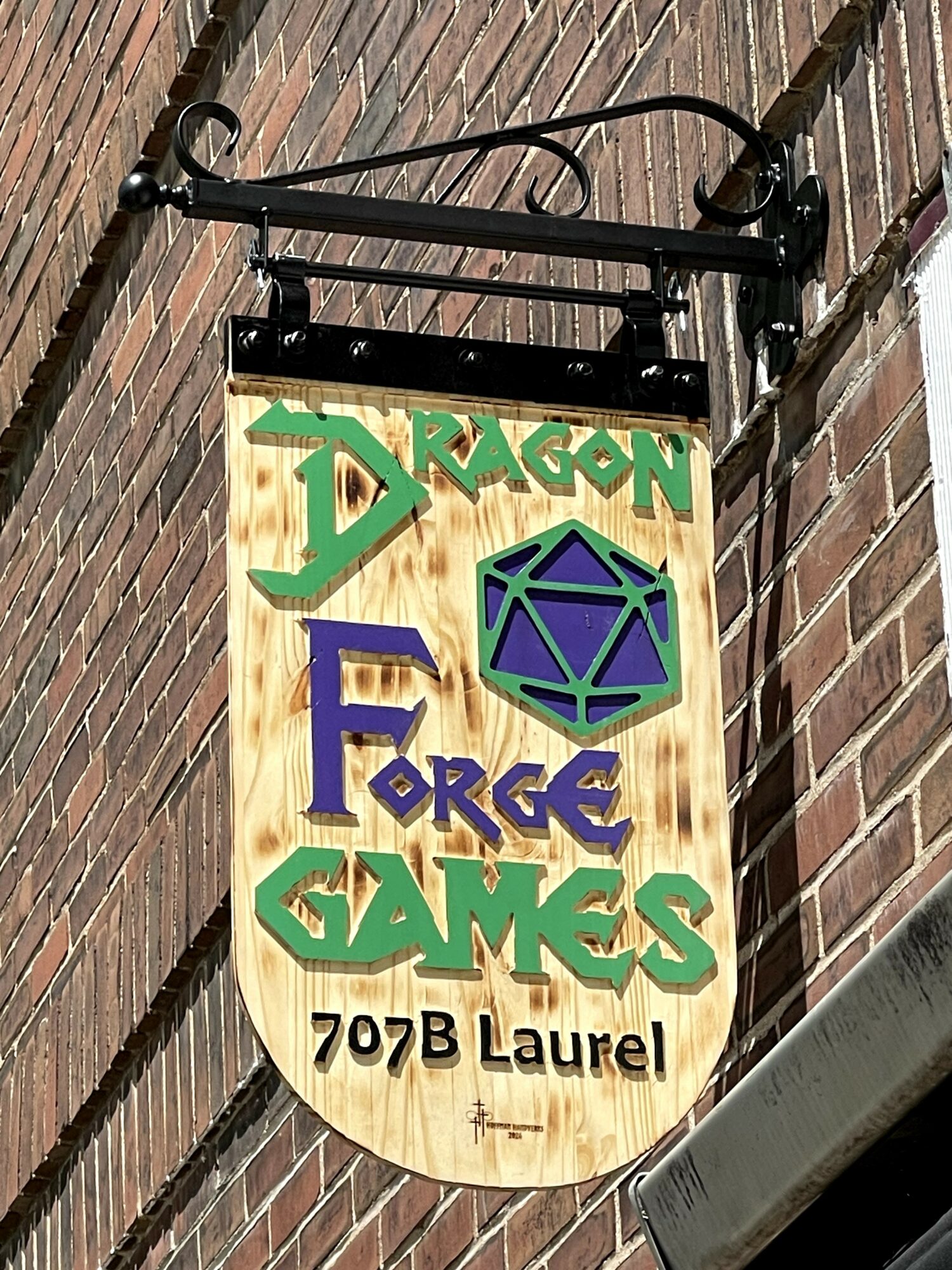 Wooden sign with green and purple text and a purple geometric shape, hanging on a brick wall.
