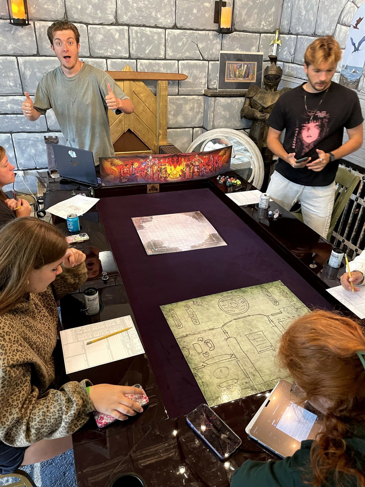 Group of people around a table with a gaming mat, some drawing, and a laptop, in a room with stone wall.
