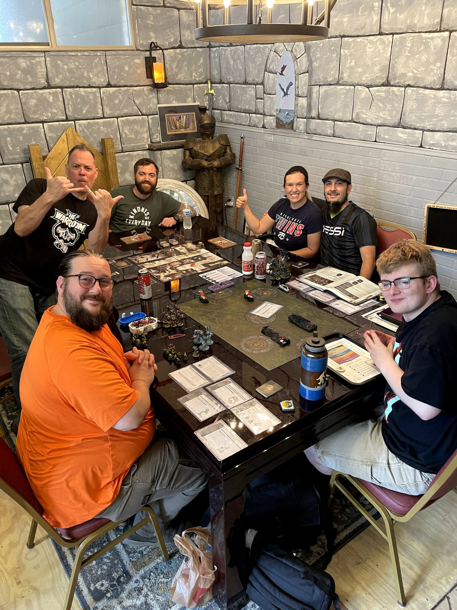 Six people gathered around a table playing a tabletop game, some giving thumbs up, in a room with stone walls and decorations.