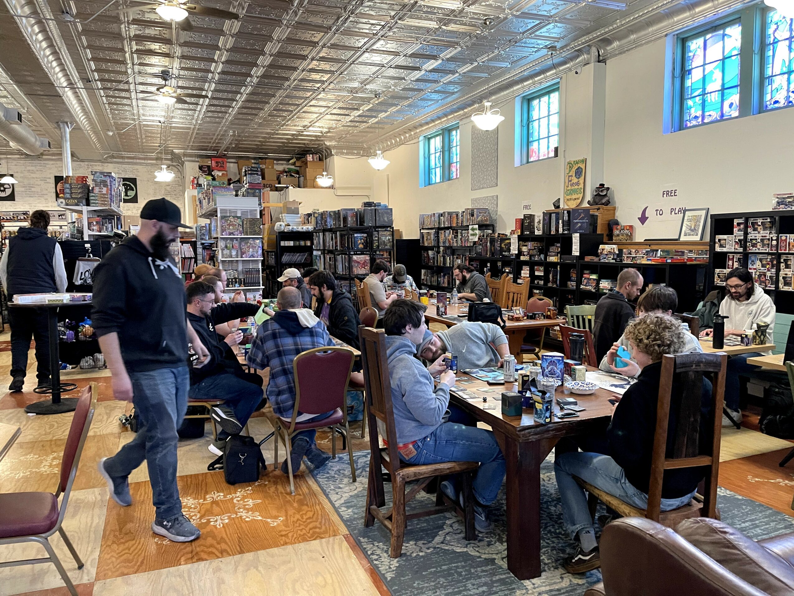 People seated at tables in a large room with bookshelves and windows, some standing and browsing books.