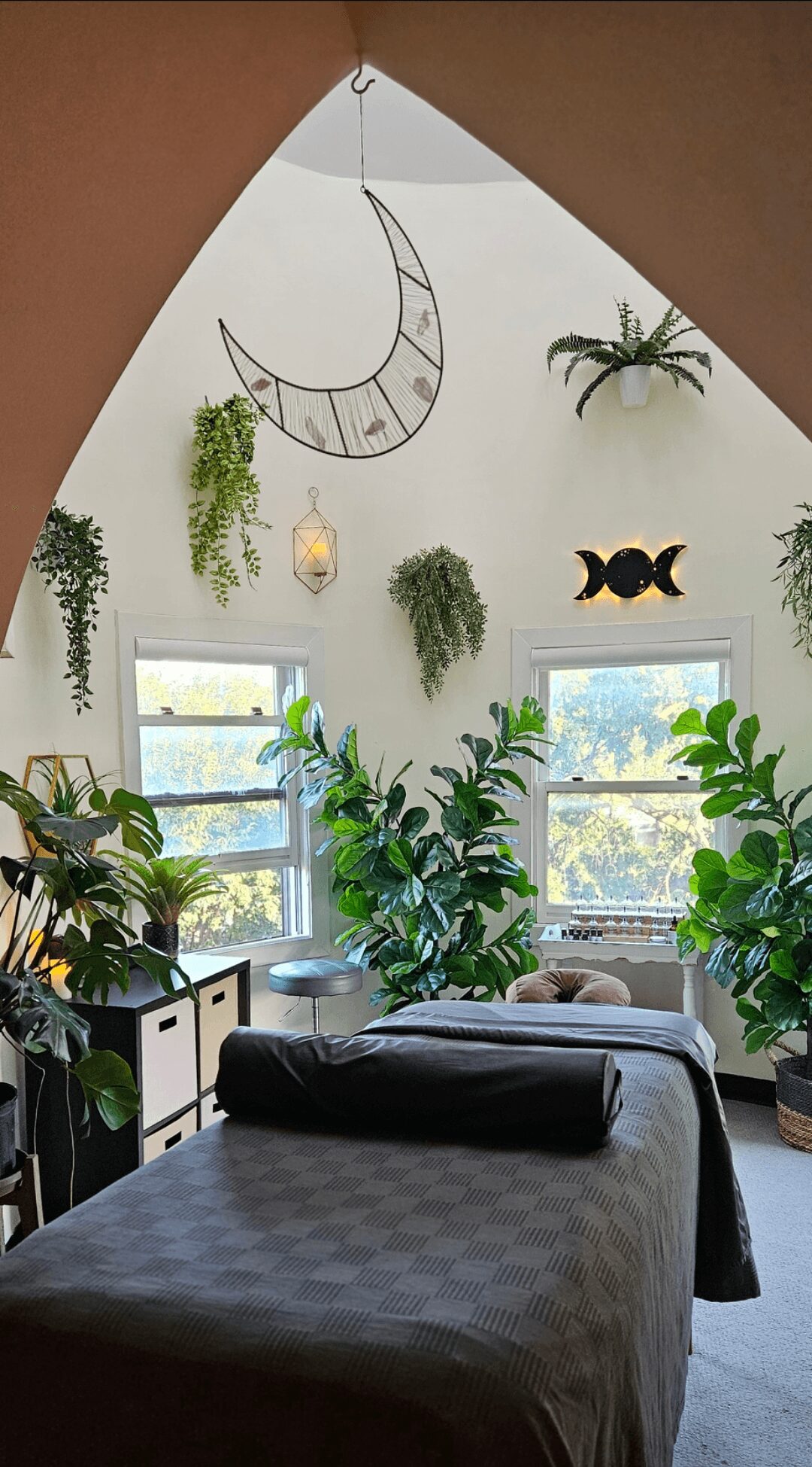 Room with large windows, green plants, hanging decor, and a bed with a rolled blanket, bright and airy atmosphere.
