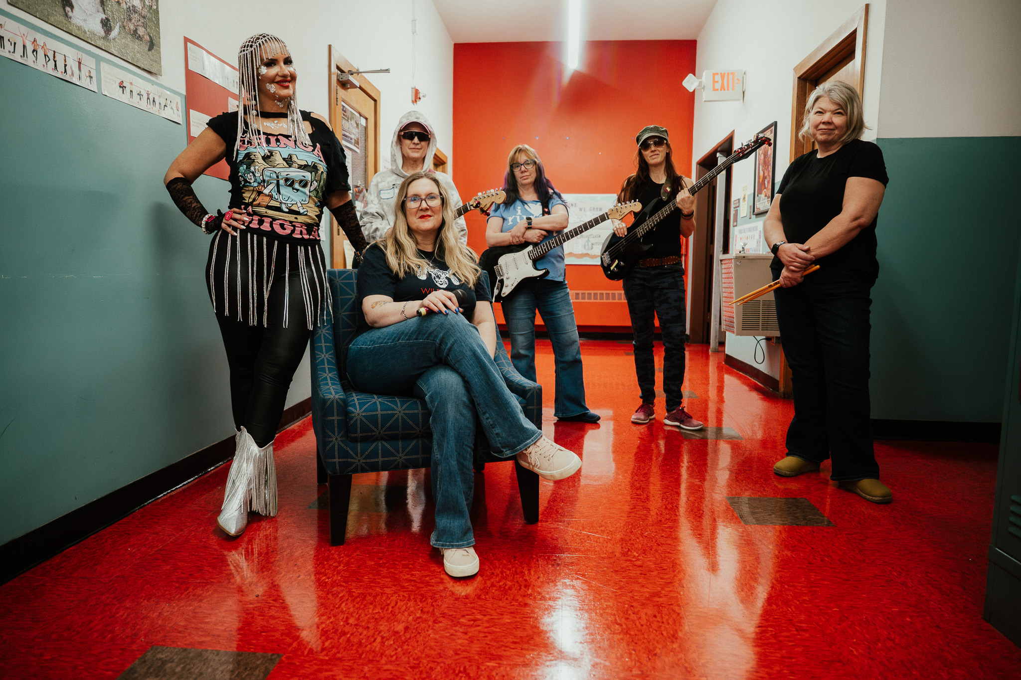 Five women in a hallway, one sitting on a chair, others standing or holding musical instruments, smiling and posing.