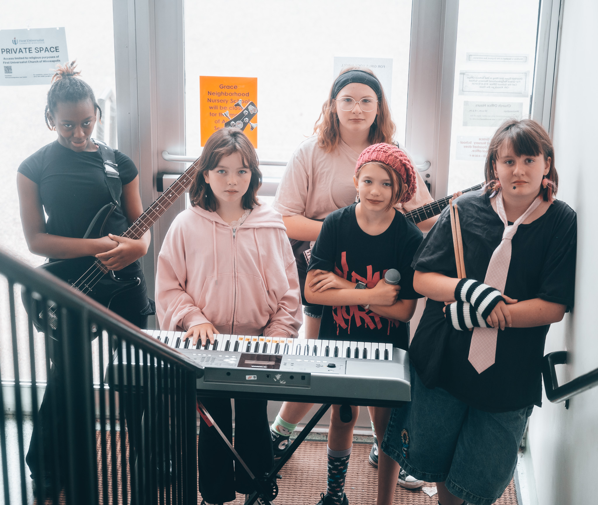 Five young girls with musical instruments and a keyboard in a room with large windows.