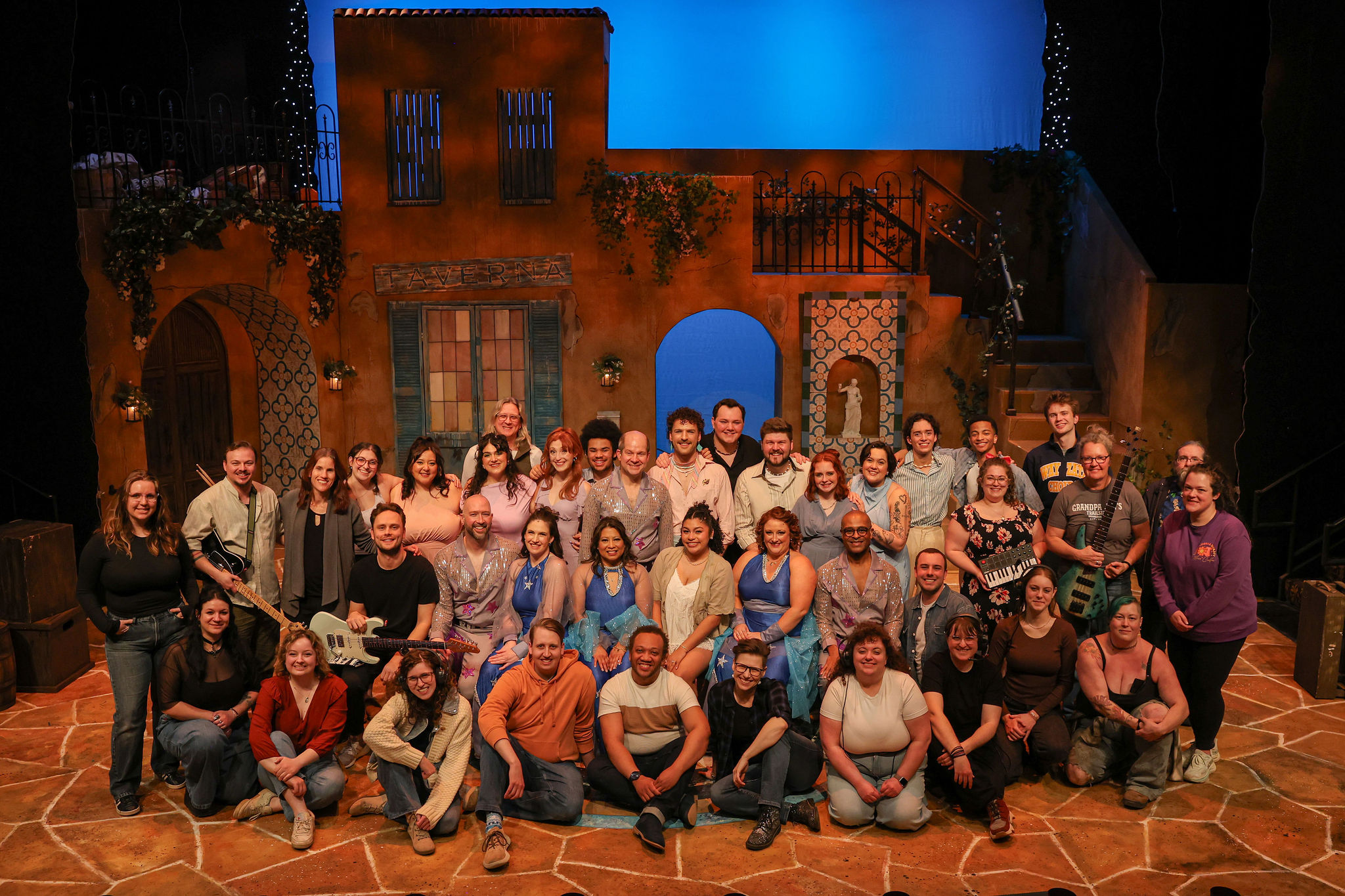 Group of people posing on stage with a backdrop of buildings and a blue sky, some holding musical instruments.