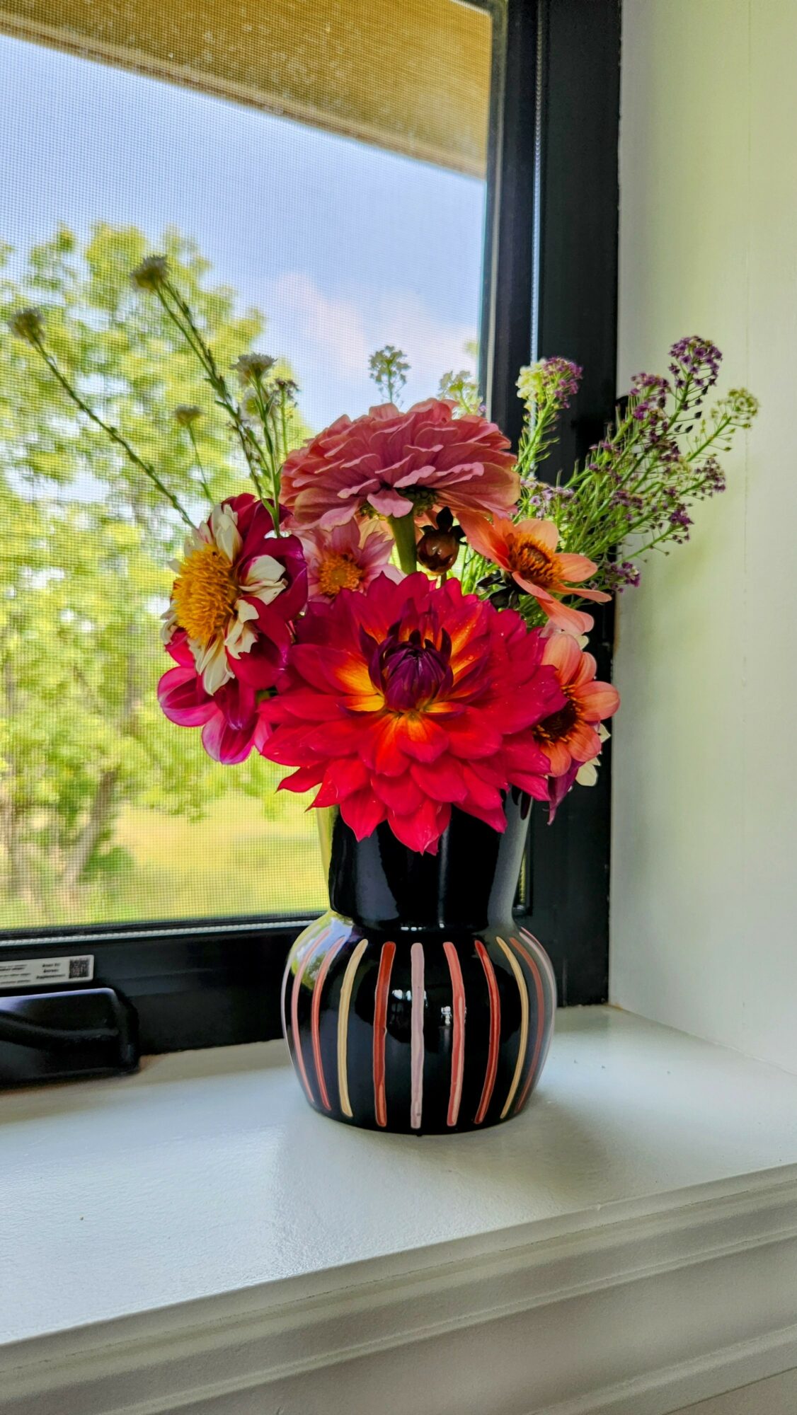 Colorful flower bouquet in a striped vase on windowsill with green trees outside.